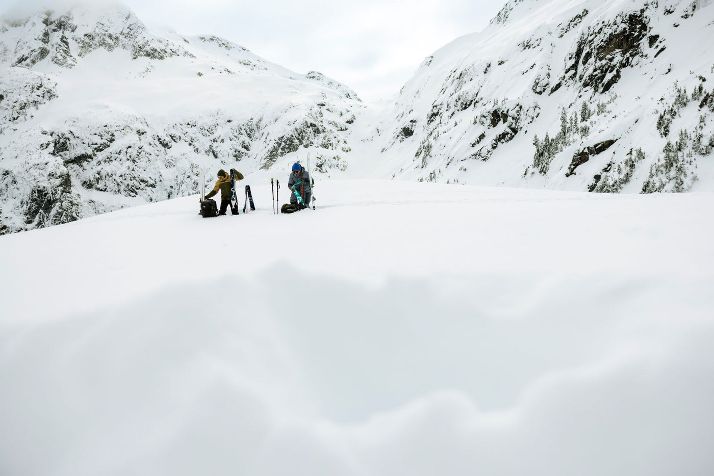 Two people with skis, climbing skins, and ski poles standing on a snowy mountain